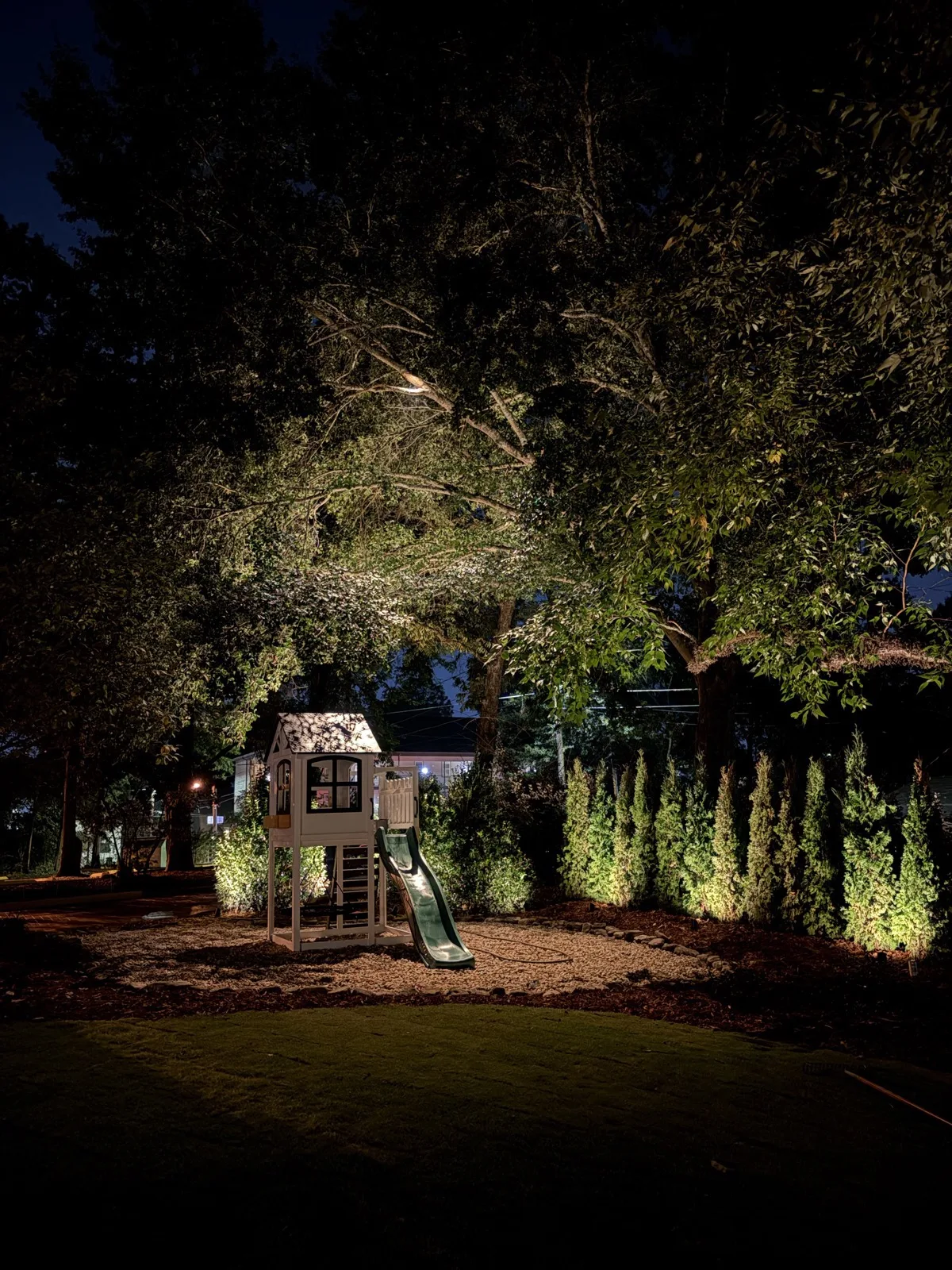 Children's playset under up-lit trees in landscaped backyard